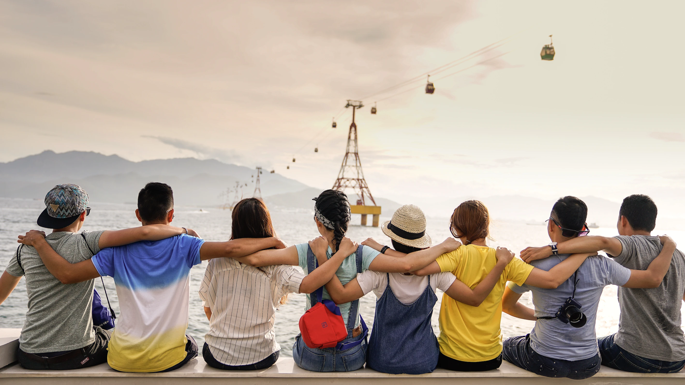 Eight children sit close with their arms over eachother, overlooking the ocean with gondolas ferrying people to a nearby island.